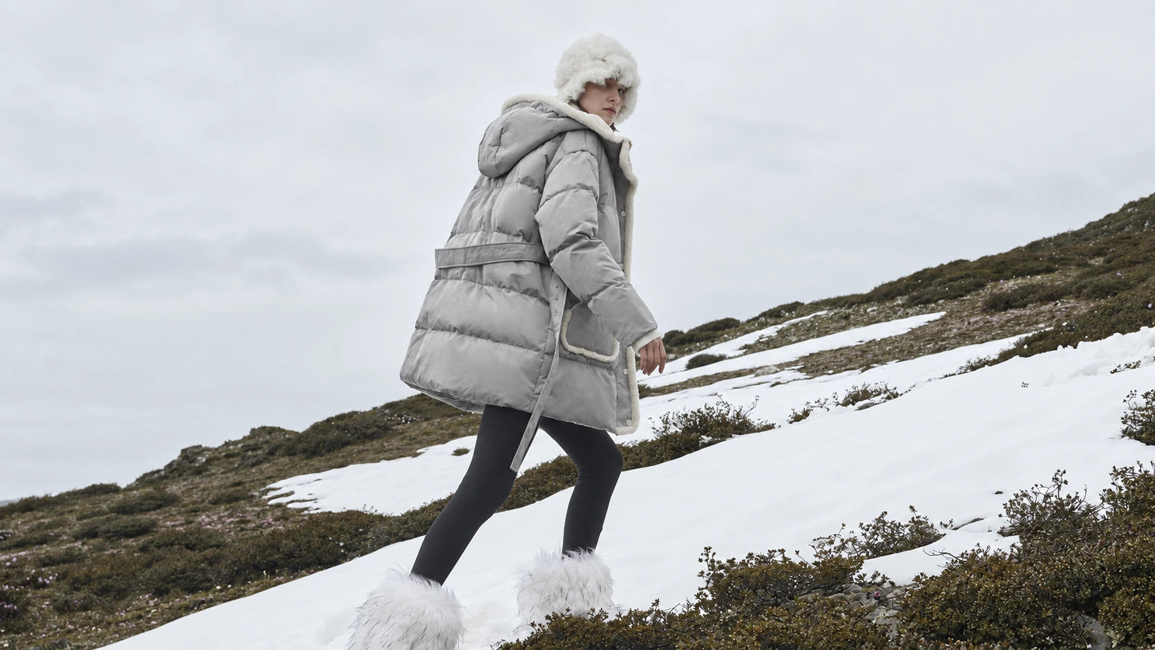 A woman in a grey belted puffer walking across a snowy hillside.